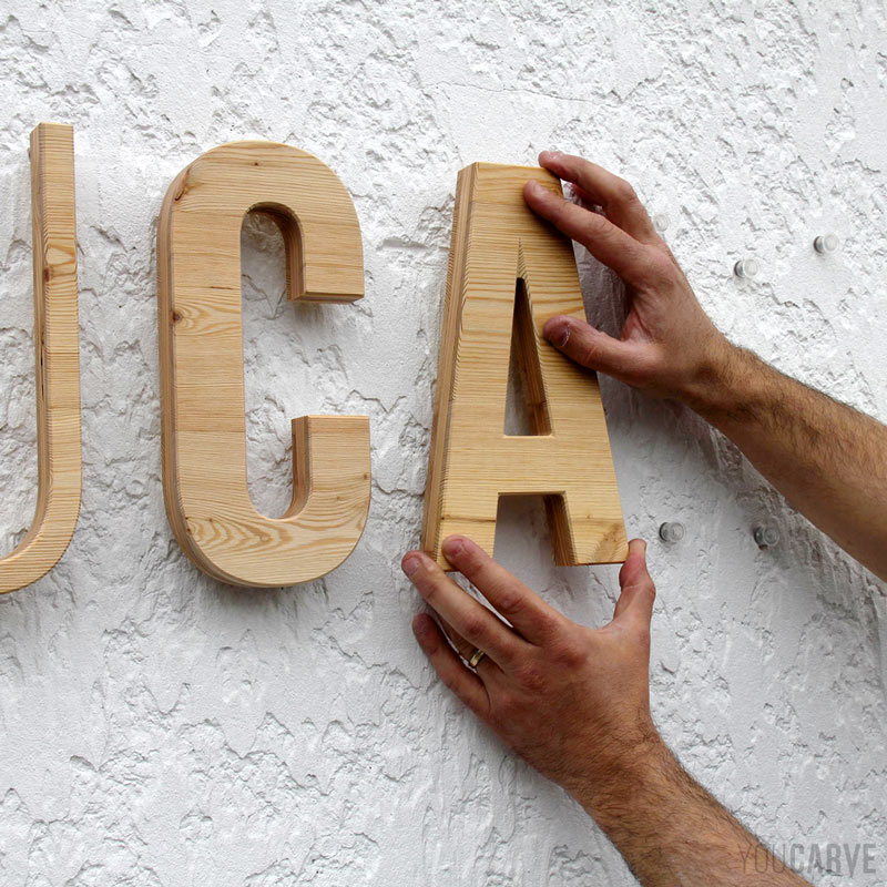 Installation enseigne - Etape 4b - Clipsage des lettres en bois Installation sur un mur d'une enseigne en lettres découpées en bois (mélèze). Gros plan sur deux mains en train de clipser la lettre "A" au mur à l'aide de fixations par entretoises emboîtables.