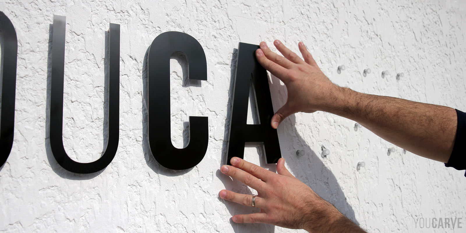 Installation enseigne - Etape 4b - Clipsage des lettres Installation sur un mur d'une enseigne en lettres découpées en alu-dibond noir. Gros plan sur deux mains en train de clipser la lettre "A" au mur à l'aide de fixations par entretoises emboîtables.