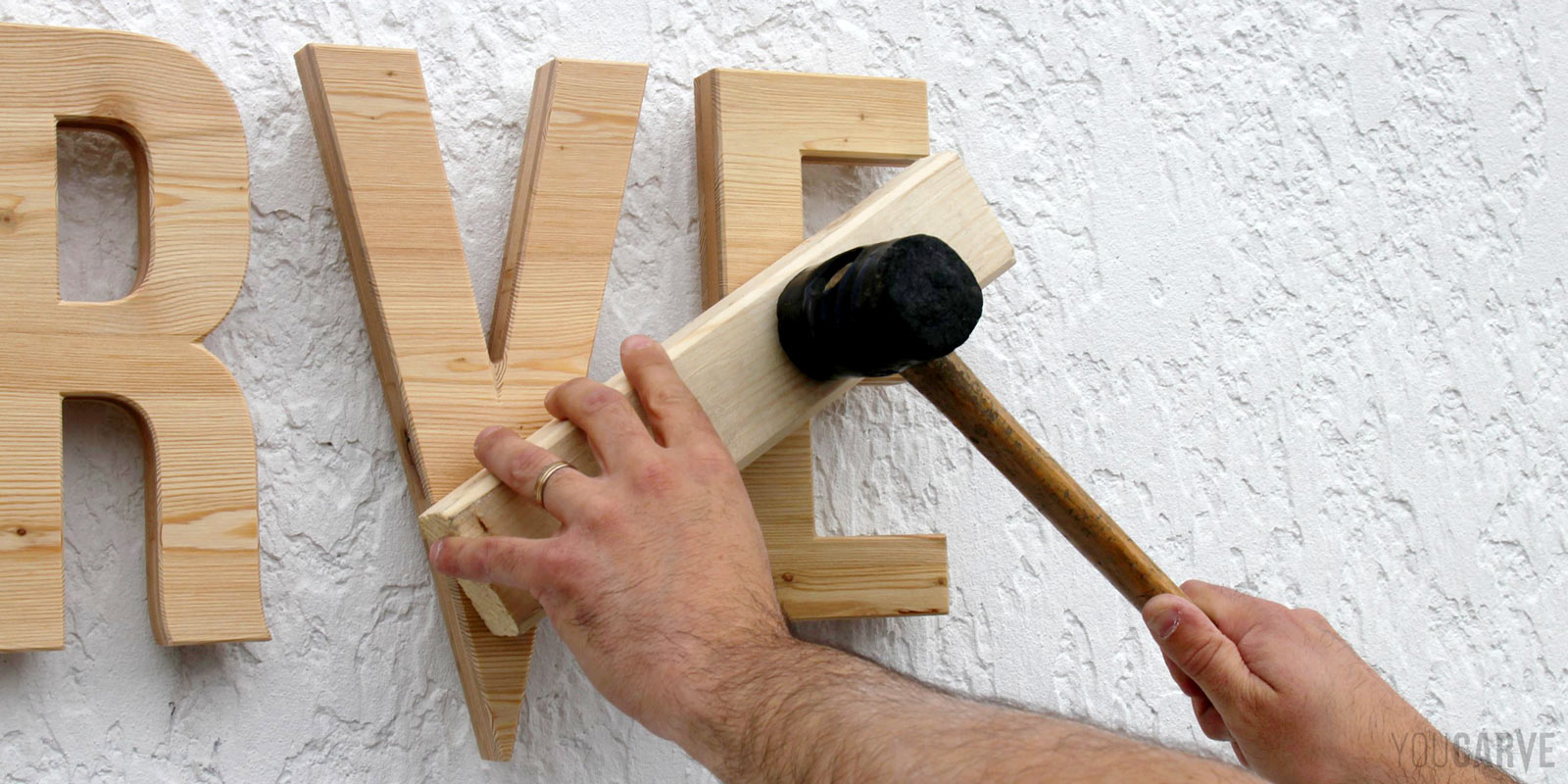 Installation enseigne - Etape 4c - Clipsage des lettres en bois Installation sur un mur d'une enseigne en lettres découpées en bois (mélèze). Gros plan sur deux mains en train de finaliser le clipsage de la fixation par entretoises à l'aide d'une cale et d'un maillet.