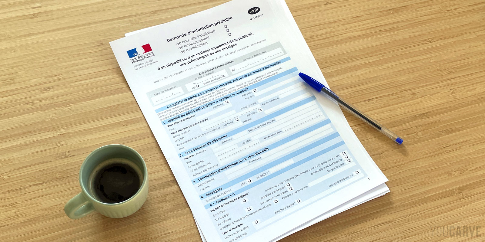 Youcarve - Article - Signage authorization request - Complete guide Blank Cerfa 14798 form (signage authorization request) placed on a wooden table with a ballpoint pen and a cup of coffee.