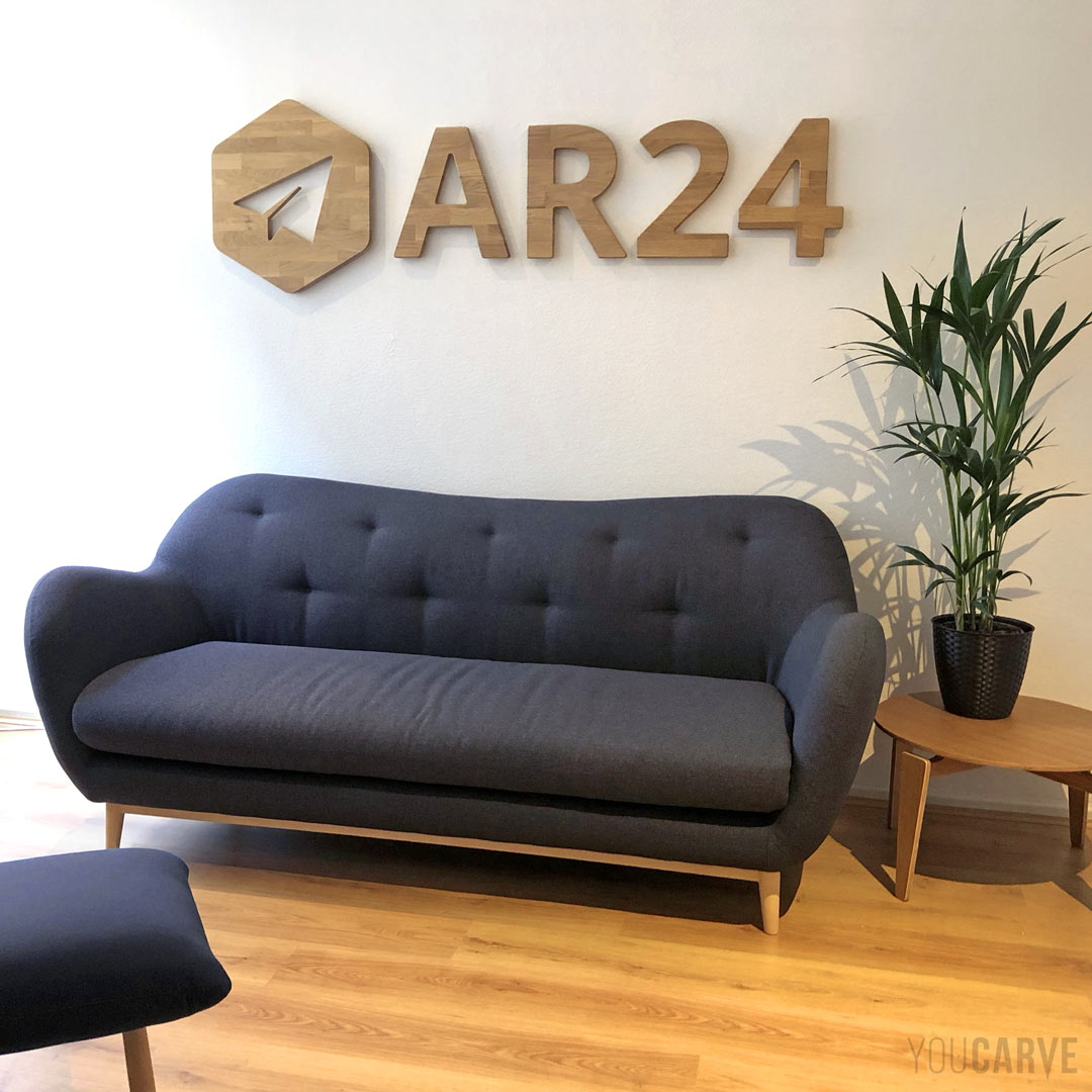 Réalisé par AR24 (recommandé électronique en ligne) : logo mural en bois, découpe sur-mesure en chêne lamellé-collé épaisseur 19 mm, fixation sur entretoises avec gabarit de perçage.