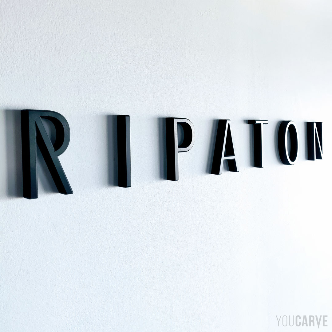 Réalisé par Ripaton (design de mobilier à Montpellier) : logo mural en relief, découpe sur-mesure en PVC expansé noir épaisseur 19 mm, fixation mousse-double-face avec gabarit de positionnement.