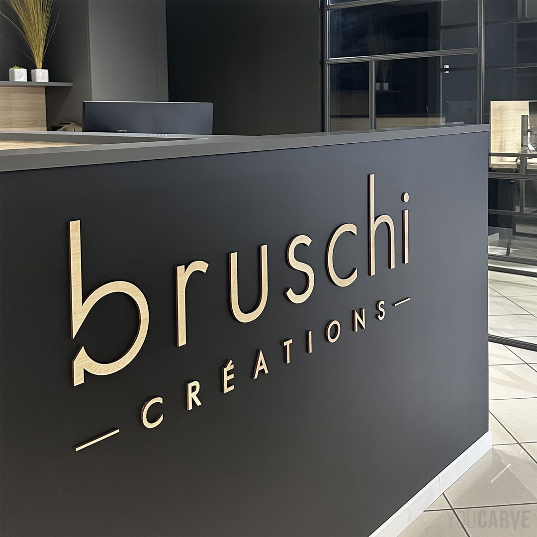 Réalisé par Bruschi Créations (cuisiniste à Reims) : logo mural en bois pour la décoration de bureau, découpe laser en érable massif épaisseur 5 mm, fixation double-face avec gabarit de positionnement.