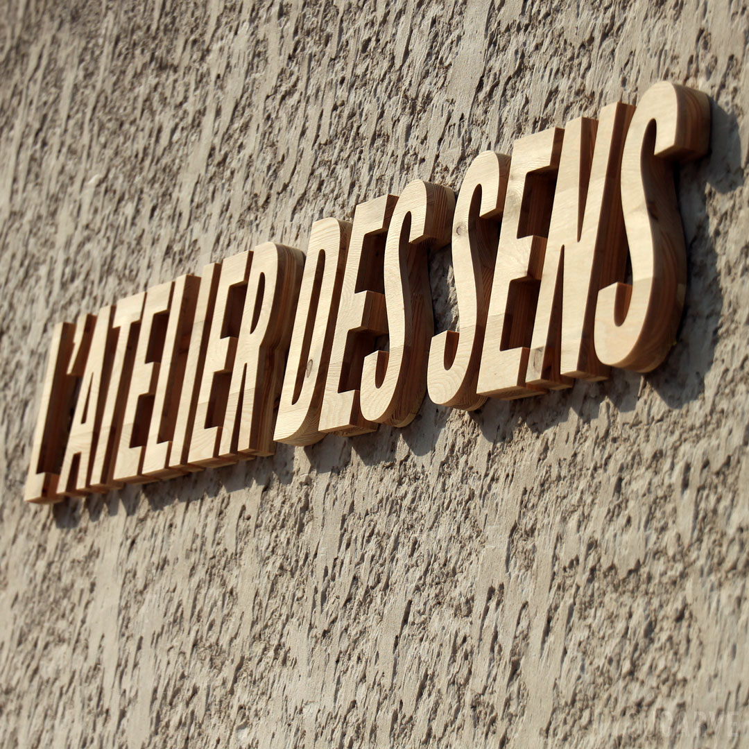 Réalisé par L’Atelier des sens : enseigne en relief, lettres découpées en bois (mélèze épaisseur 26 mm), fixation sur entretoises avec gabarit de perçage.