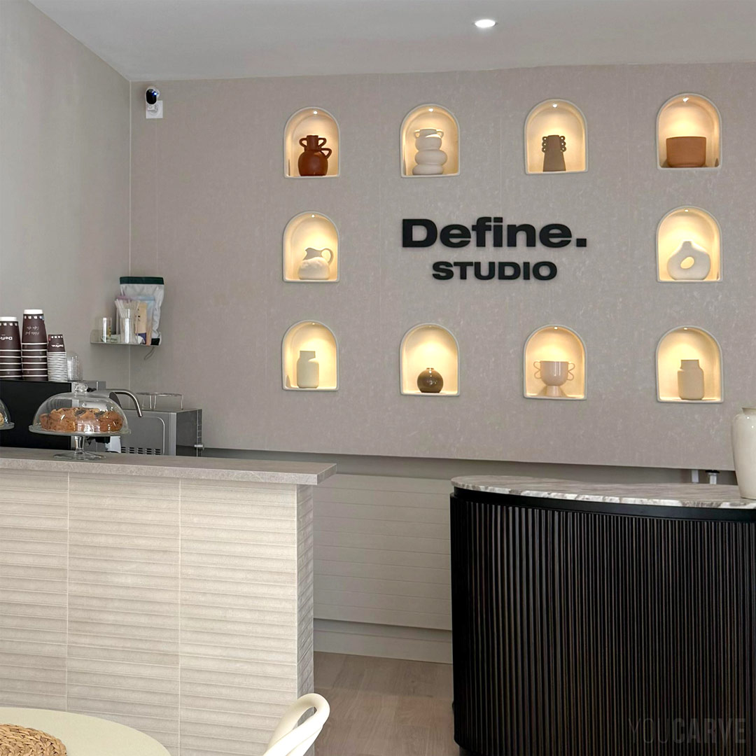 Define. Studio Nice - Logo mural en relief (PVC expansé noir ép. 19 mm) Enseigne murale intérieure "Define. Studio" à l’accueil, lettres découpées en PVC expansé noir, fixation mousse double-face sur un mur beige avec des niches décorées.