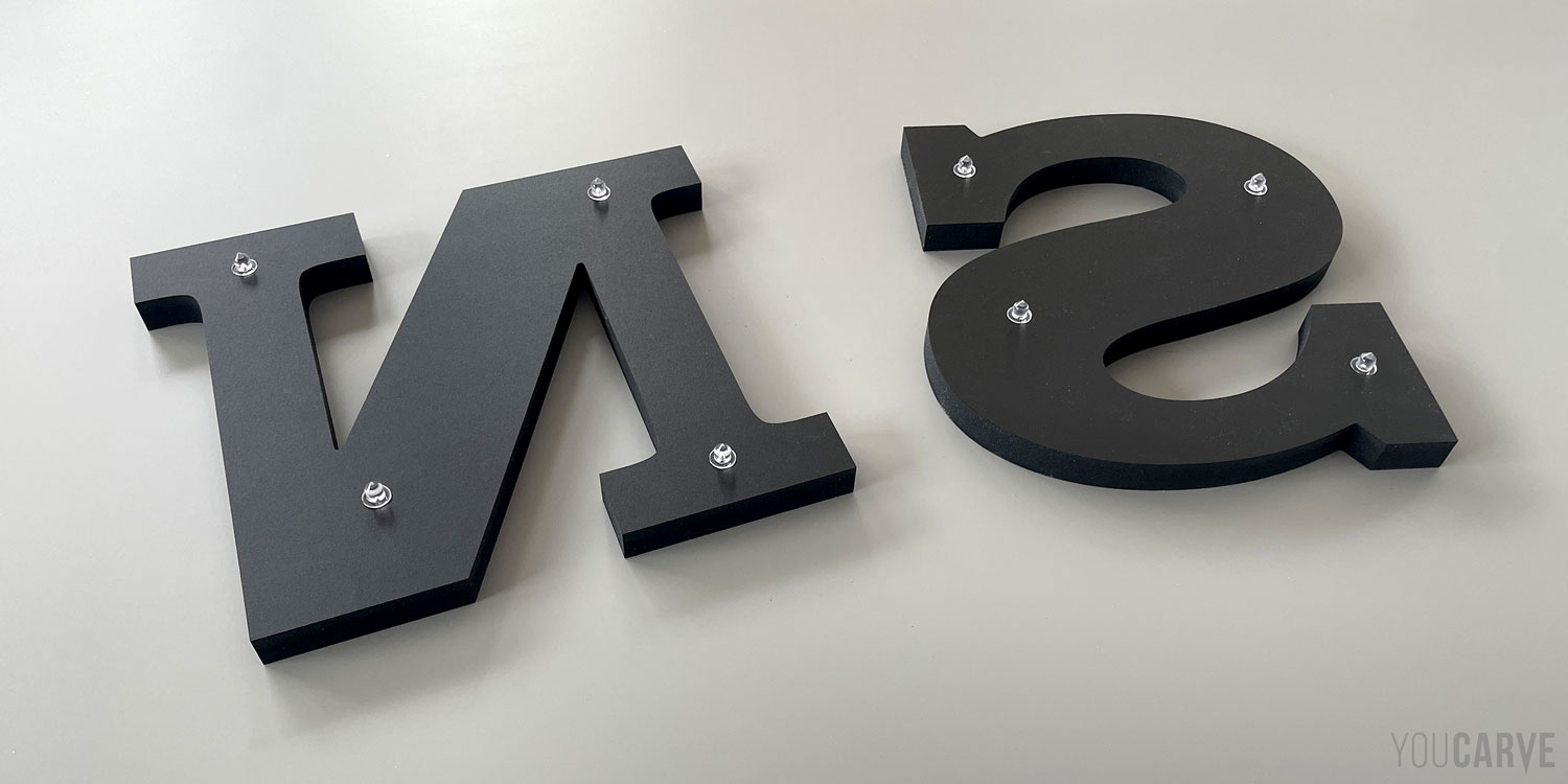 Fixation par entretoises avec effet détrompeur Vue sur du verso de deux lettres d’enseignes (N et S) en PVC expansé noir, avec leurs fixations (parties mâles) collées aux dos et positionnées asymétriquement