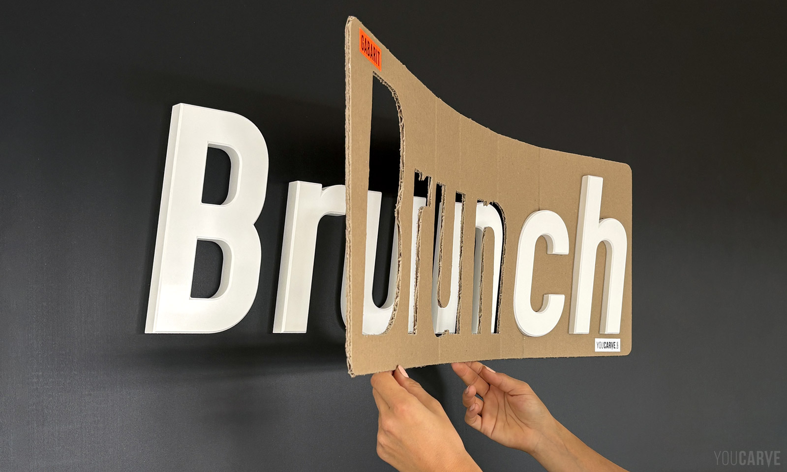 Youcarve - Article - Gabarits de pose, l’atout décisif pour installer lettres d’enseigne et logos muraux Pose d’un lettrage mural 'Brunch' en lettres découpées en PVC blanc sur un mur gris anthracite. Deux mains retirent le gabarit de pose en carton, révélant les lettres déjà fixées et parfaitement alignées.
