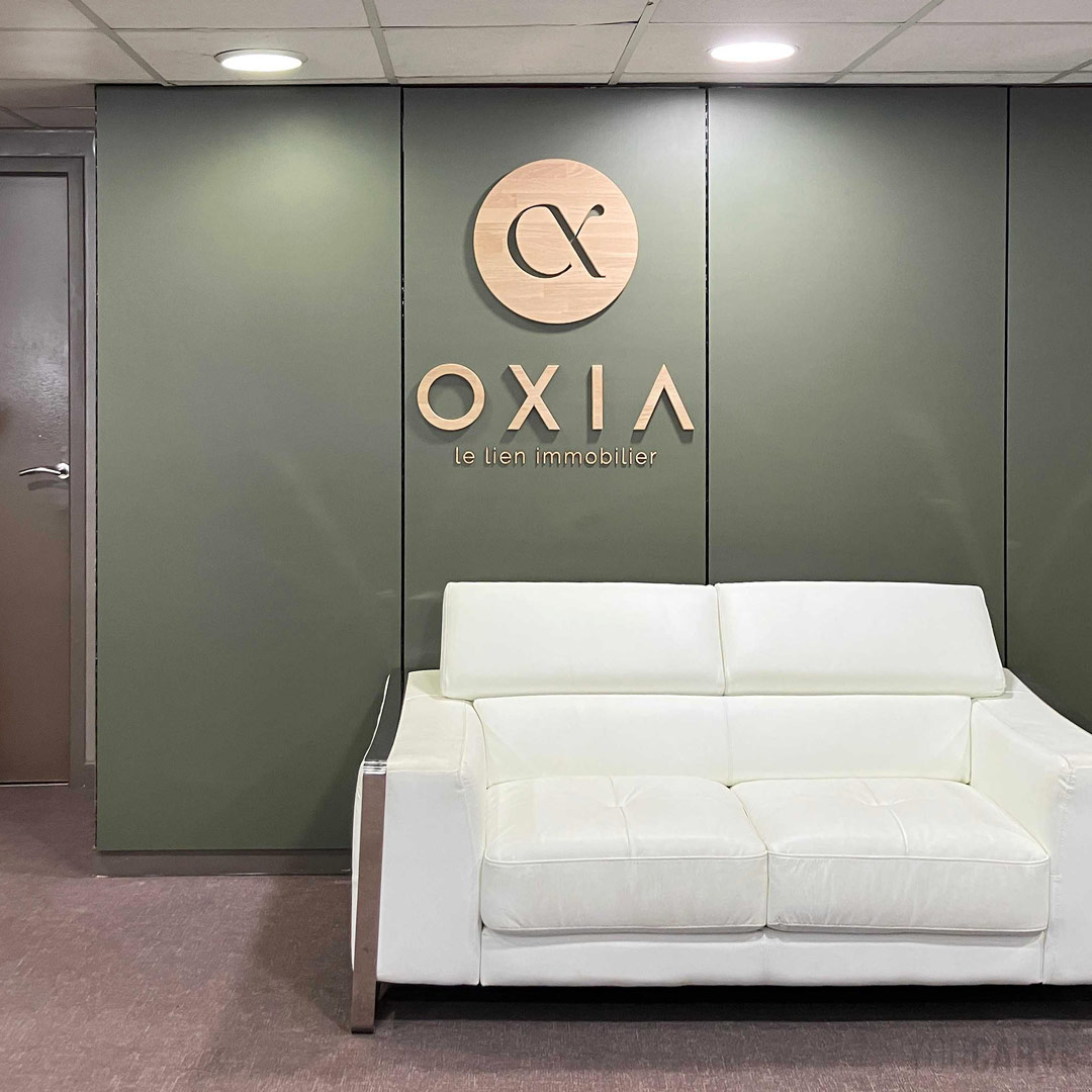 Oxia Lyon - Logo mural en bois (chêne 19 & 5 mm) Logo mural Oxia en bois (chêne naturel) fixé sur une cloison vert sauge derrière un canapé blanc