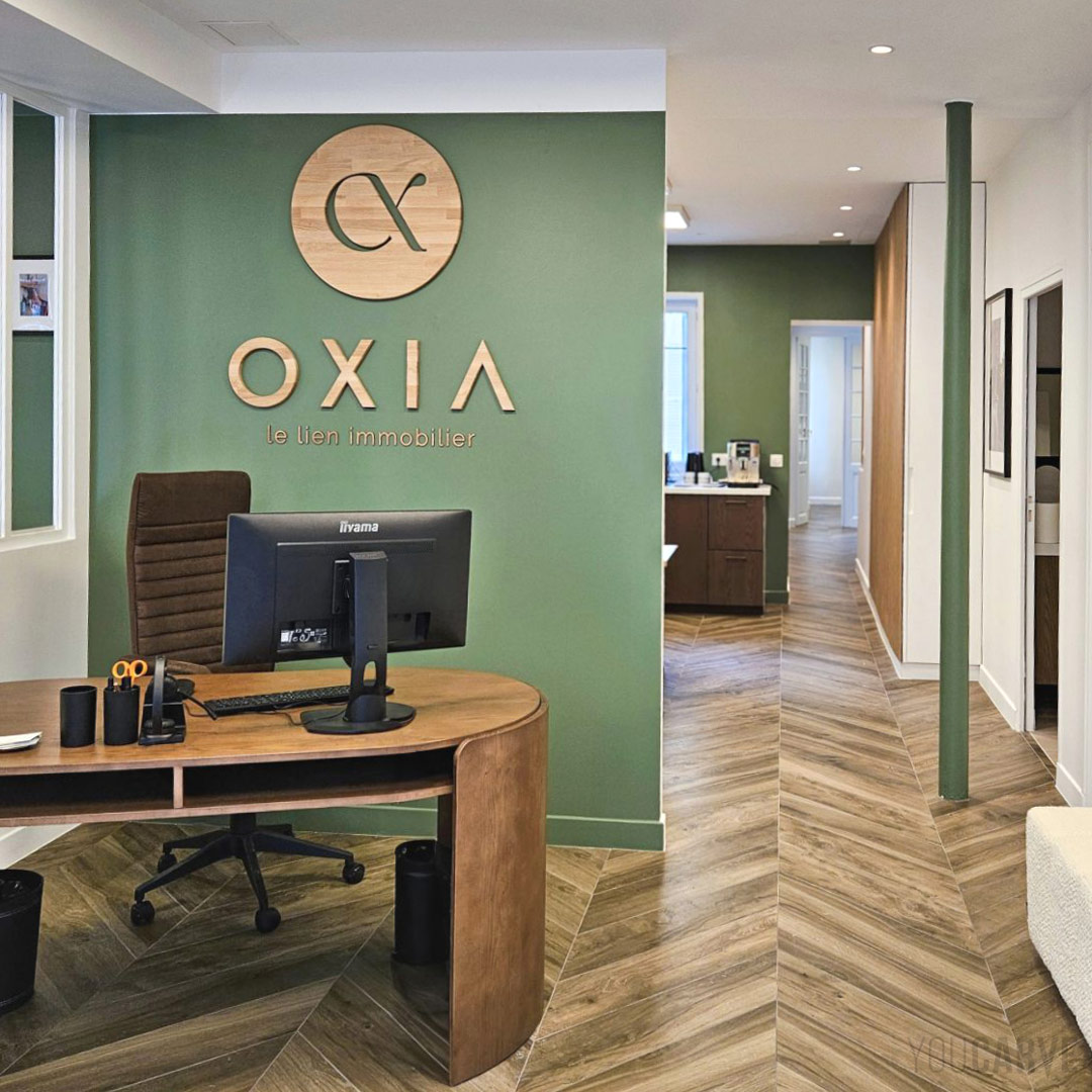 Oxia Paris Montparnasse - Logo mural en bois (chêne 19 & 5 mm) Logo mural Oxia en bois (chêne naturel) fixé sur une cloison vert amande derrière un bureau en bois